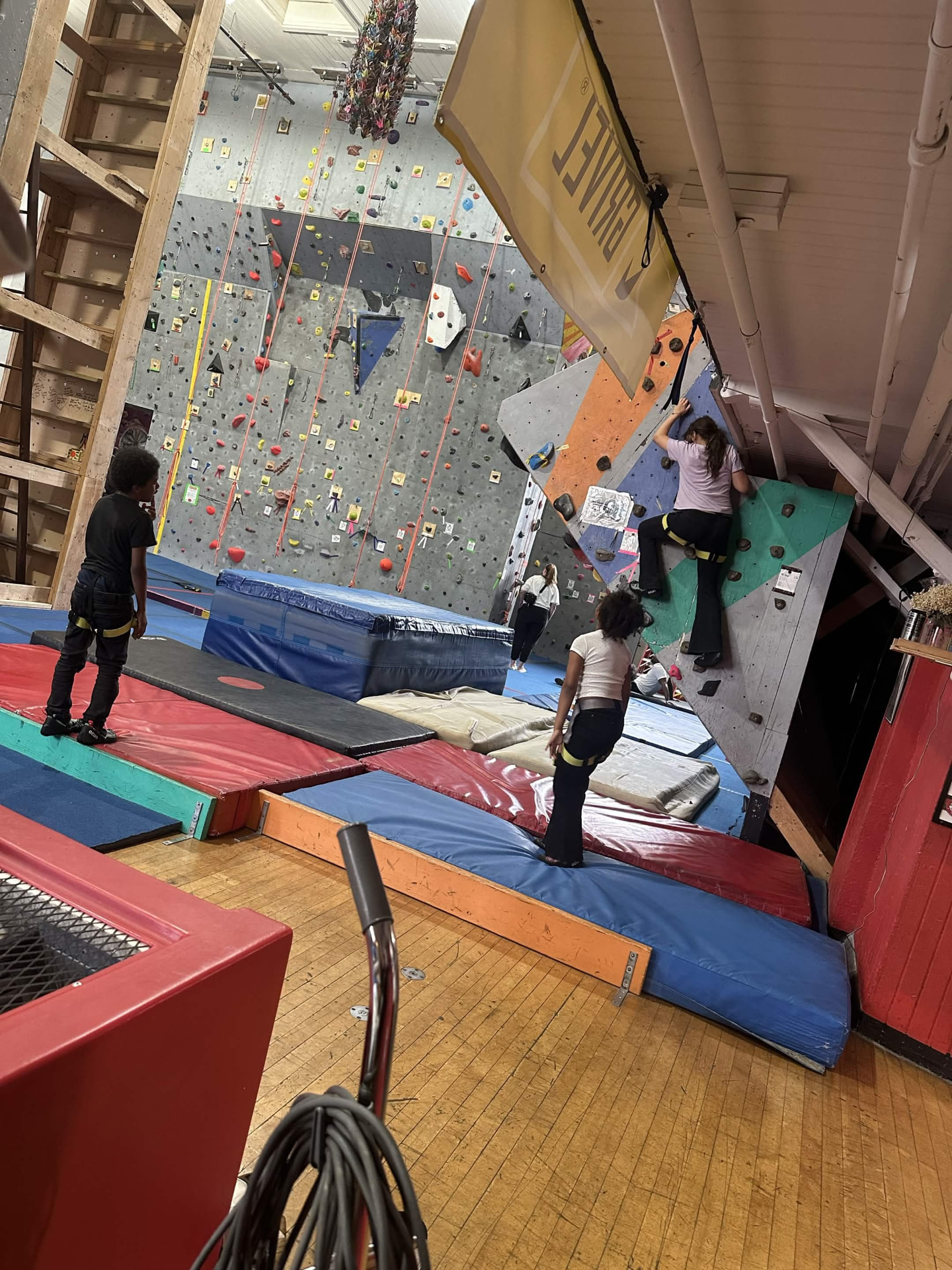 A young person climbing a bouldering wall while peers watch and encourage.